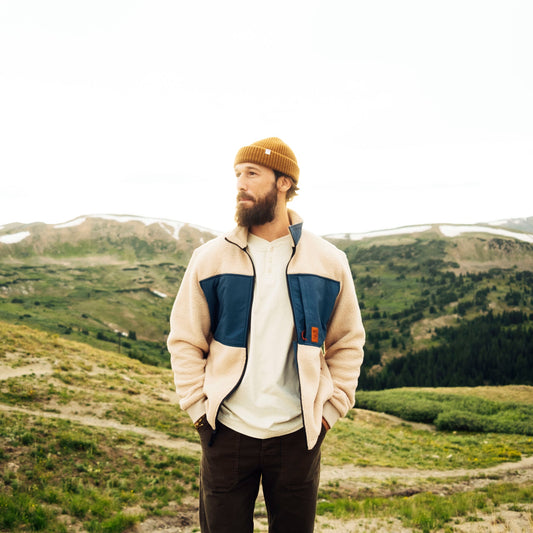 Man standing in a mountainous landscape wearing a beige jacket with blue accents and a brown beanie.