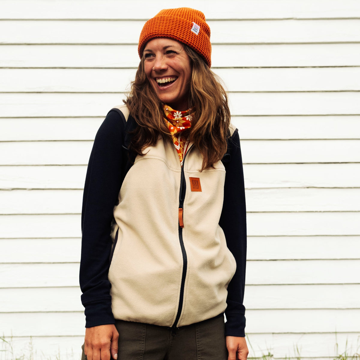 Person wearing a beige and black jacket with an orange beanie and scarf, standing against a white wall.